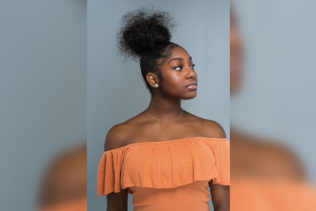 Jovem com coque volumoso e baby hair modelado, vestindo blusa ombro a ombro em tom laranja