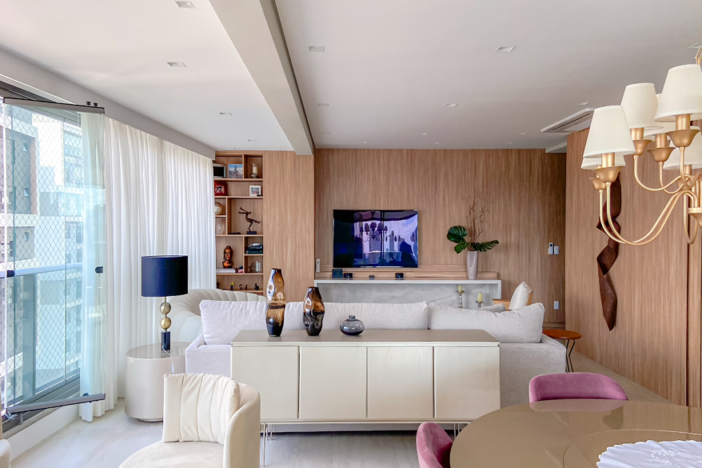 Sala de estar com móveis claros e buffet bege encostado nas costas do sofá com vasos em cima e itens de decoração, com uma parede de vidro e as demais em madeira, e a mesa da sala de jantar é bege escura com duas cadeiras estofadas em roxo e uma poltrona branca