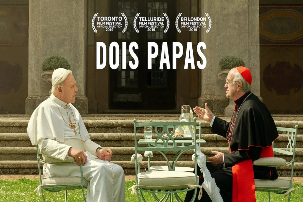 Dois homens sentados conversando em jardim, um deles está usando uma roupa litúrgica branca e o outro com uma roupa litúrgica preta e vermelha