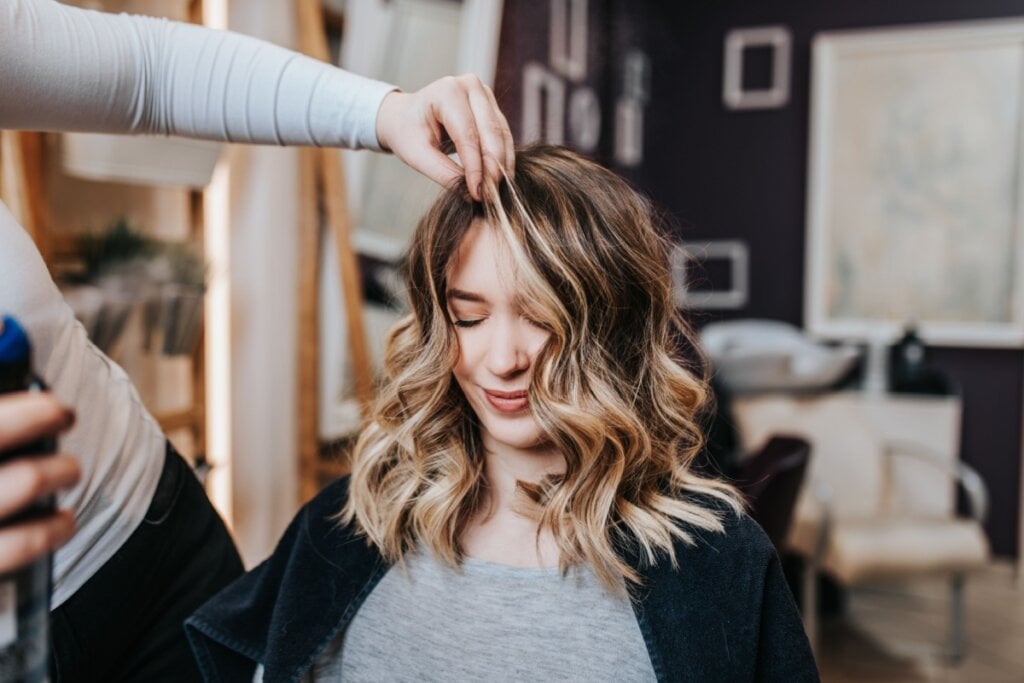 Mulher com cabelo com mechas loiras sentada e uma mão mexendo em seu cabelo 