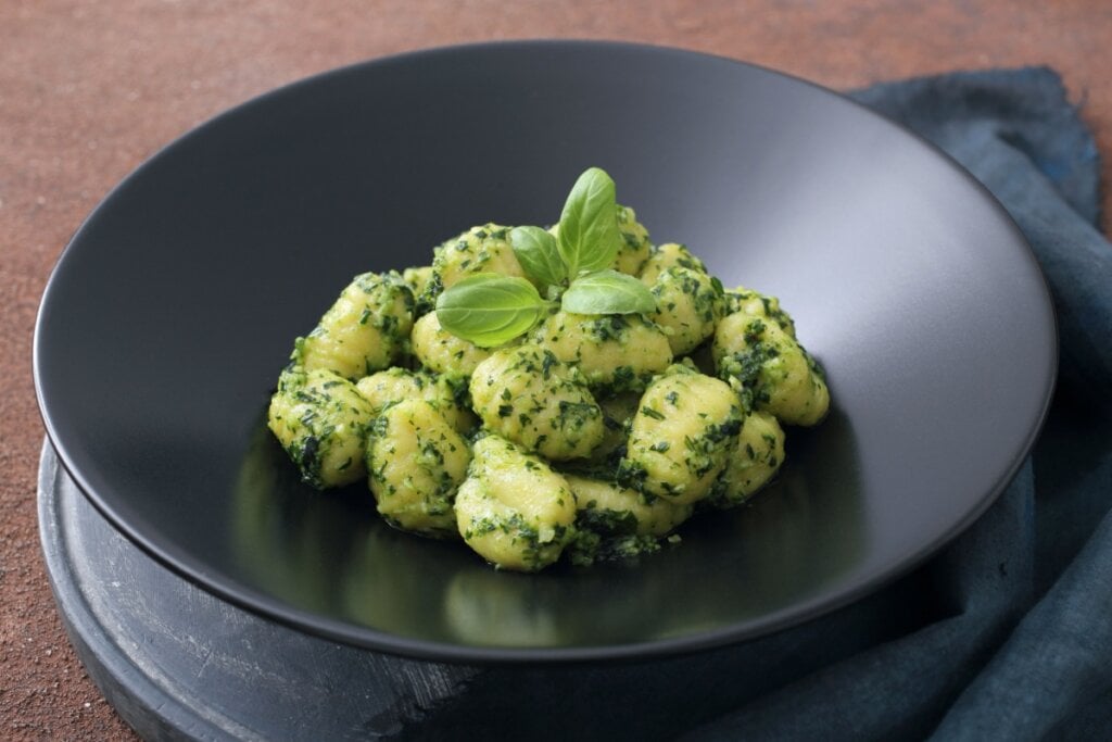 Nhoque ao molho pesto servido em prato fundo preto em cima de mesa de madeira