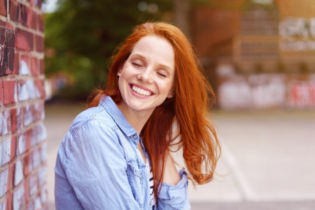 Mulher com cabelos ruivos acobreados sentada na rua e sorrindo com os olhos fechados
