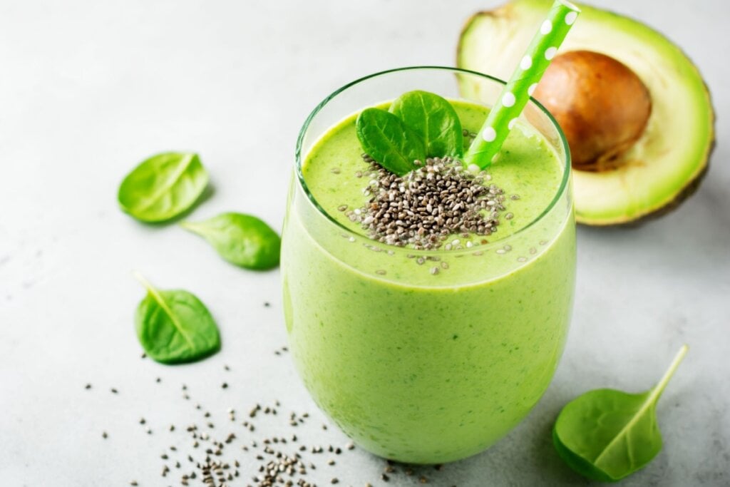 Vitamina de abacate com espinafre e chia servido em copo de vidro decorado com folhas de espinafre e chia e ao lado está um abacate cortado ao meio 