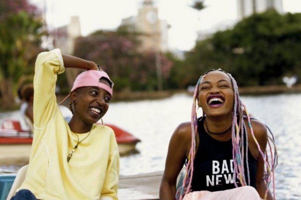 Duas jovens rindo sentadas uma ao lado da outra, uma está usando um boné rosa e uma blusa amarela e a outra tem o cabelo trançado na cor rosa e está usando uma regata preta