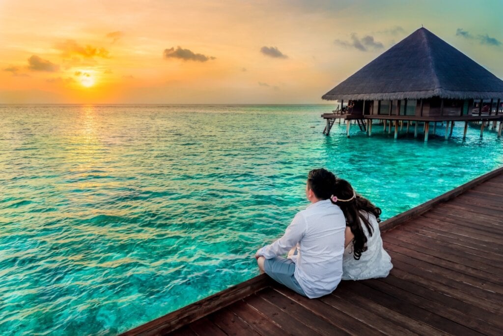 Casal sentado em um deck de madeira assistindo ao pôr do sol diante do mar azul-turquesa nas Maldivas ao lado de bangalô
