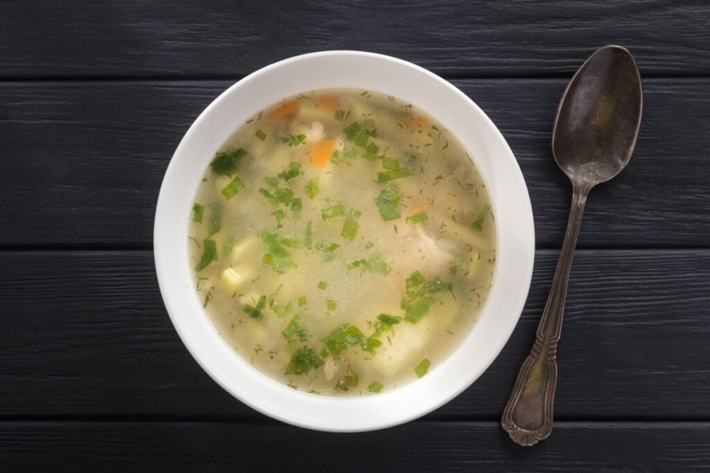 Caldo de frango servido em tigela de cerâmica branca, com colher ao lado em cima de mesa de madeira
