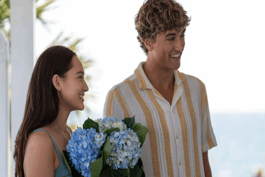 Imagem de uma jovem com o cabelo solto, usando um vestido de alcinhas verdes e com flores azuis nas mãos, ao lado um jovem com cabelo cacheado loiro, usando uma camisa listrada branca e laranja