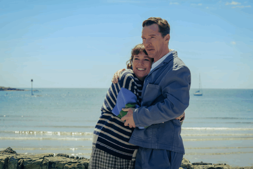 Imagem de um casal abraçado a beira do mar, ela está vestindo uma blusa listrada colorida em azul, branco, e verde e uma calça xadrez e ele um blusa jeans azul e calça jeans azul 