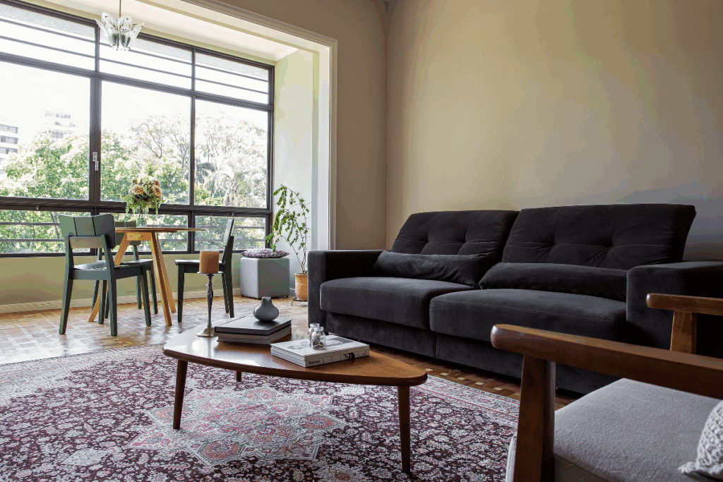 Sala de estar com sofá preto, tapete estampado e amplas janelas de vidro e mesa de centro em madeira