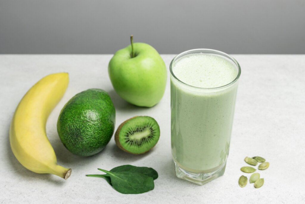 Copo de vidro com vitamina de espinafre, banana, maçã, abacate e kiwi, sobre uma mesa decorada com sementes de abóbora