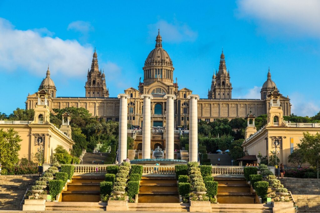 Imagem da entrada do Museu Nacional d’Art de Catalunya (MNAC) com escadas e plantas nas laterais e 4 pilastras e prédio ao fundo