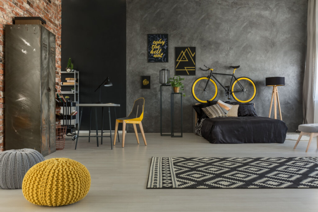Quarto com parede cinza, cama com lençol preto, tapete geométrico e puffs em amarelo e cinza