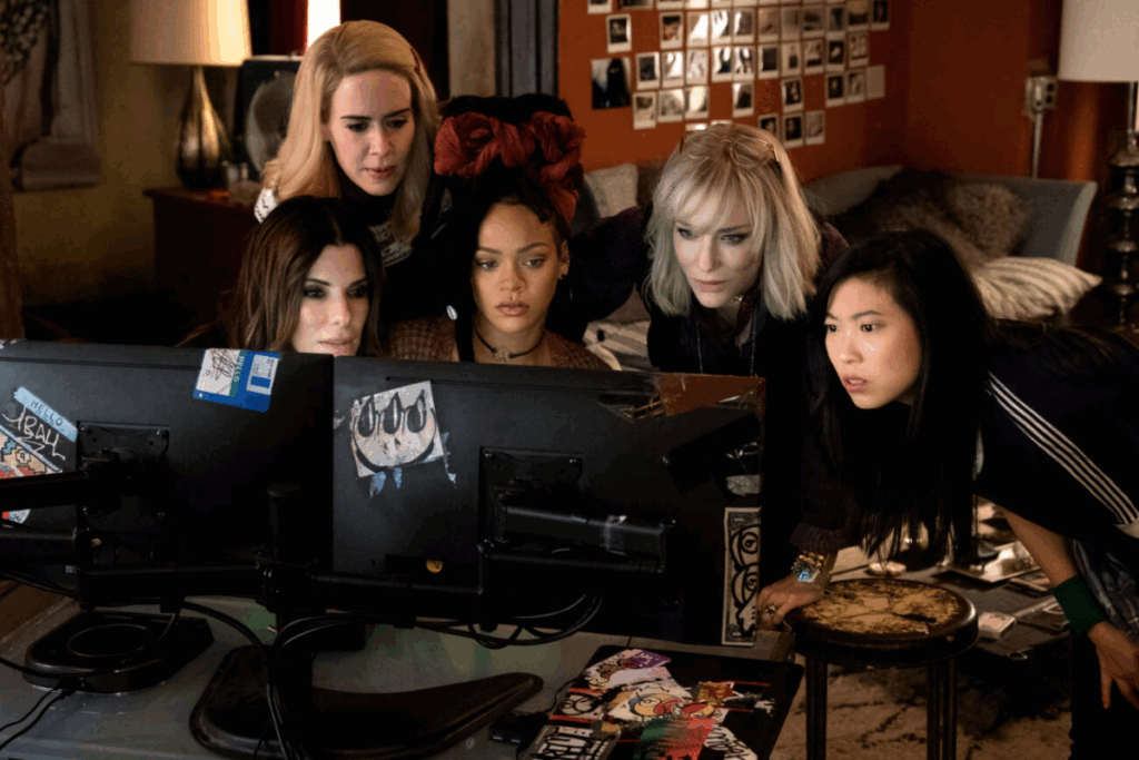 5 mulheres sentadas em frente a um computador, duas tem o cabelo loiro curto e as outras o cabelo preto e longo