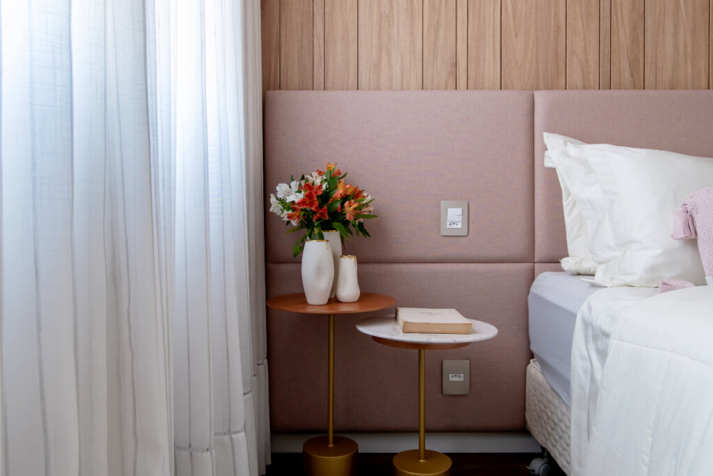 imagem parcial de quarto com cabeceira de cama rosa, duas mesas de cabeceira, uma com livro e outro com vasos brancos com flores