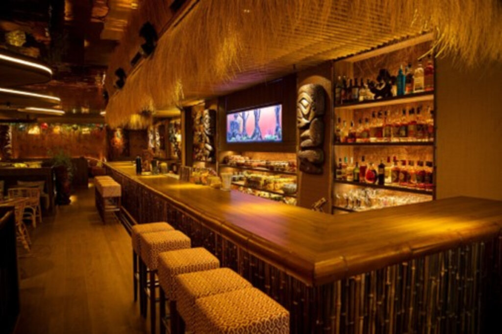 imagem de bar com luz amarela fraca com balcão grande de madeira e parede com prateleiras com bebidas. bancos em frente ao balcão