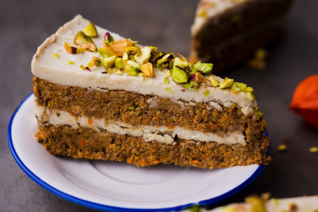 Fatia de bolo de cenoura com pistache e creme de castanha-de-caju servido em prato branco com borda azul em cima de mesa de madeira
