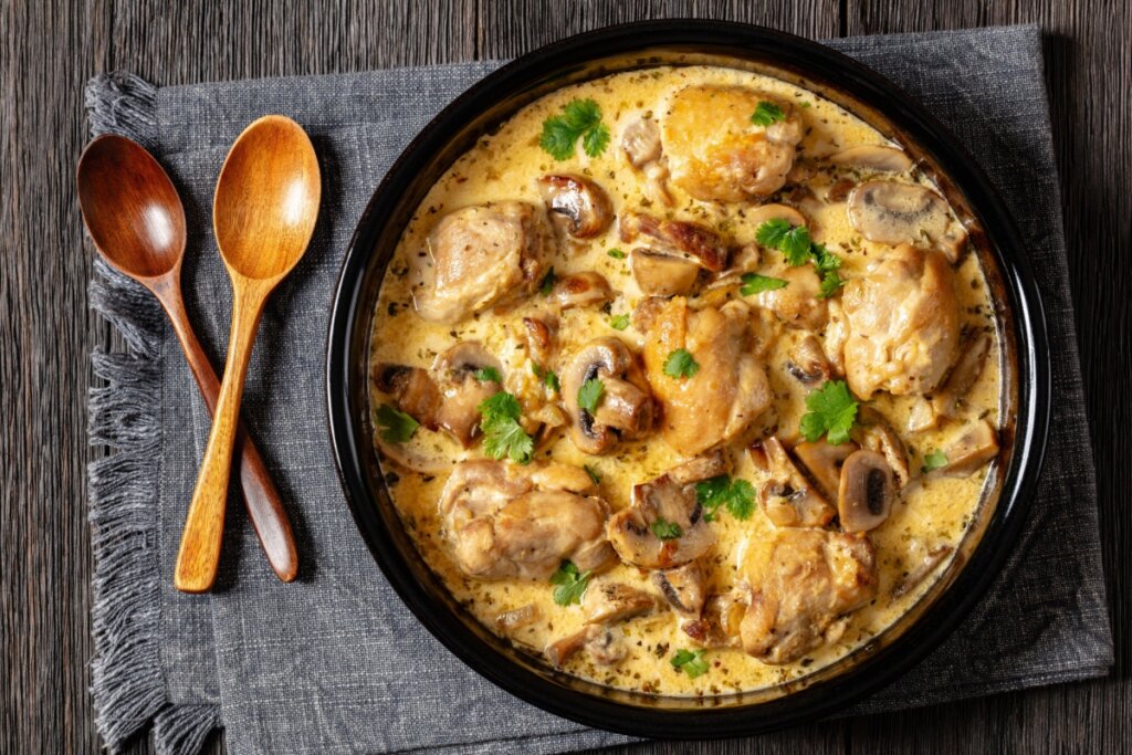 Frango ao molho de cogumelos servido em panela de ferro em cima de mesa de madeira com colheres de madeira ao lado