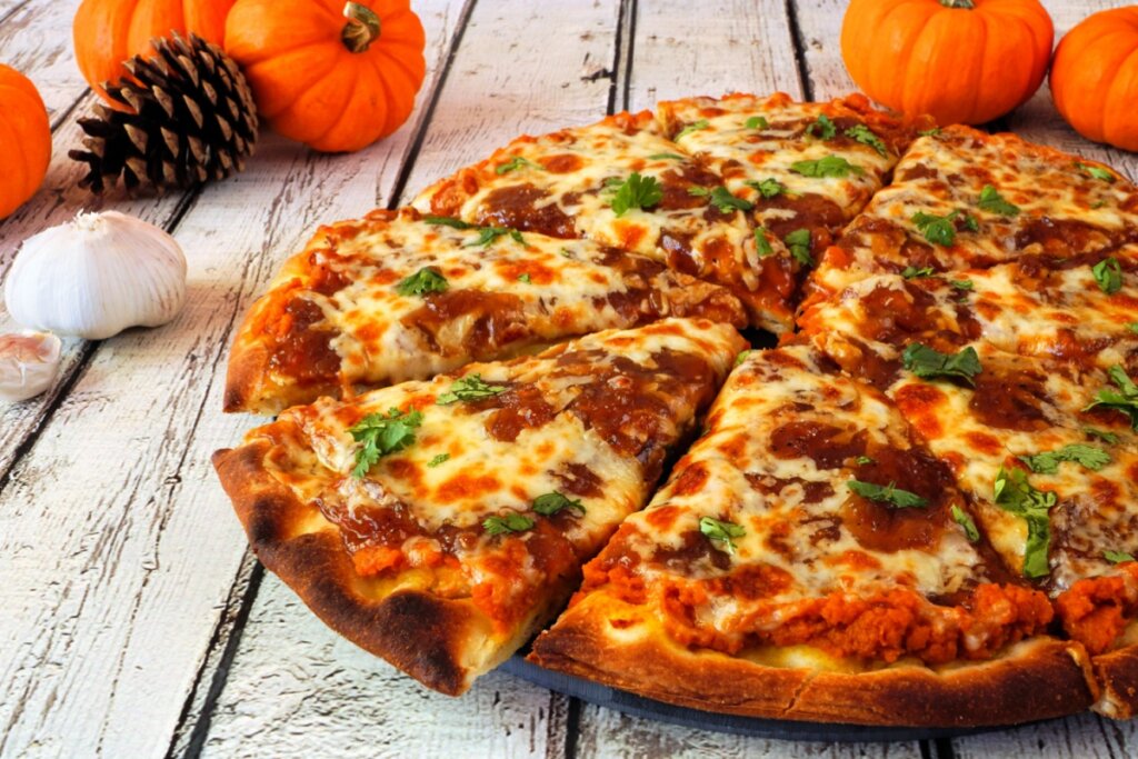 imagem de pizza com cobertura de purê de abóbora e queijo derretido em superfície de madeiras como pequenas abóboras ao fundo