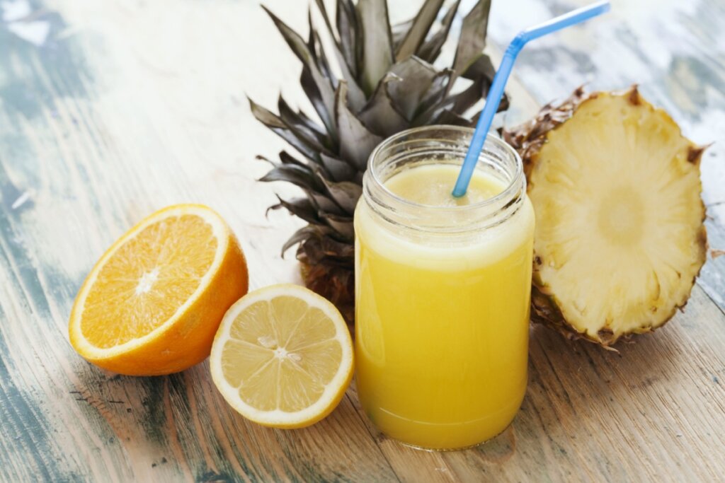 Suco de abacaxi com laranja e limão servido em copo de vidro com canudo azul, ao lado um abacaxi cortado ao meio, e uma laranja e um limão -siciliano cortado ao meio
