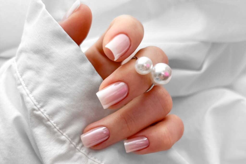 Mulher com as unhas pintadas com esmalte perolado e usando um anel com duas perolas e uma camisa branca em fundo branco