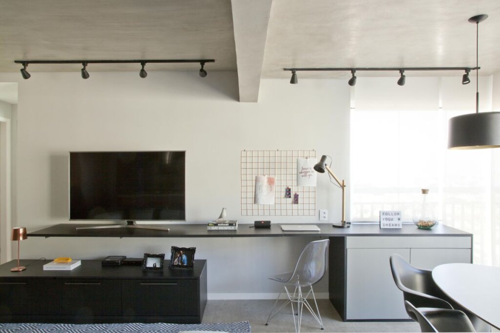 Sala de estar com televisão integrada com o home office com mesa cinza e parede brancas 