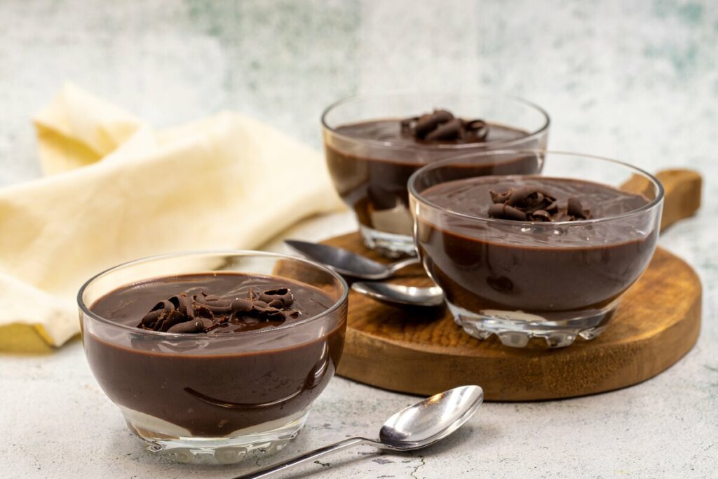 três recipientes de vidro com musse de chocolate. dois deles estão em cima de tábua. colheres ao lado