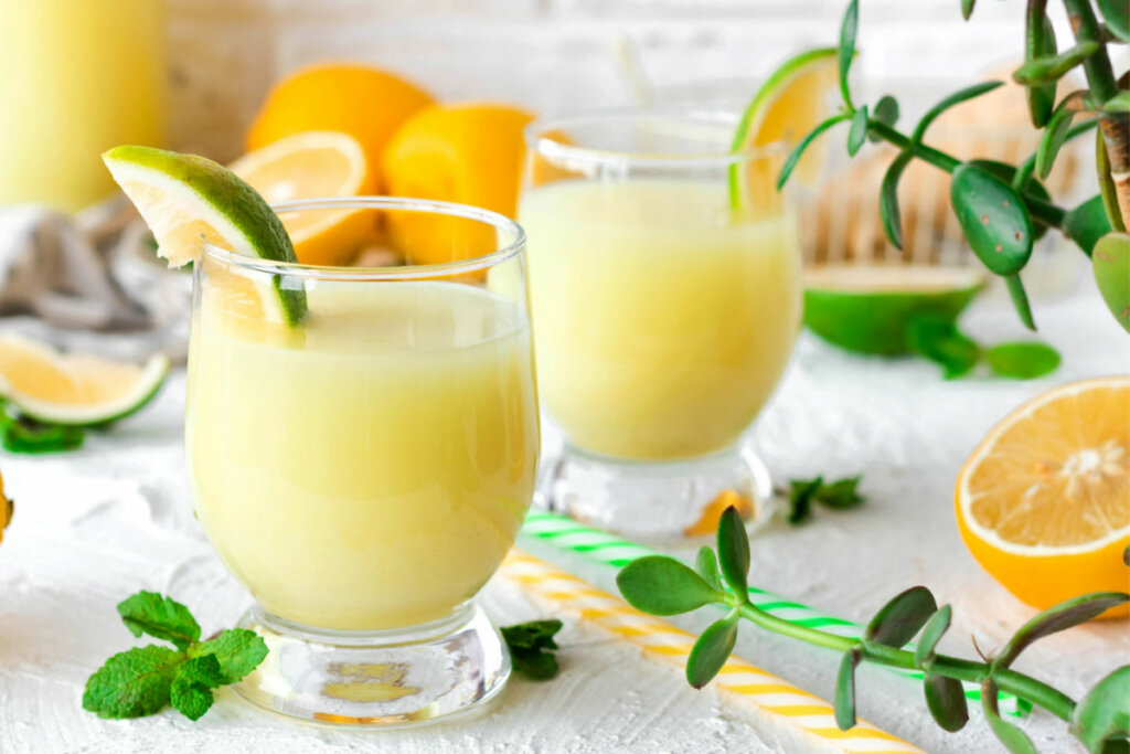 Suco amarelo em copo de vidro com rodela de laranja na borda e frutas, plantas e canudos em volta