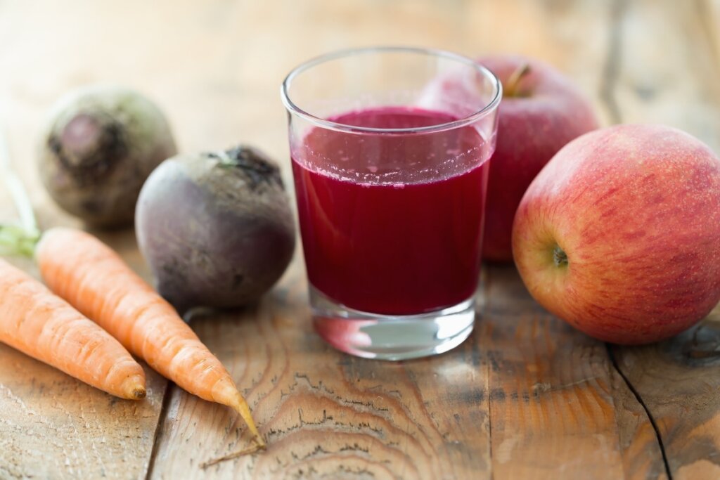 Copo de vidro com suco de beterraba com maçã e cenoura em cima de uma mesa de madeira com beterrabas, cenouras e maçãs ao redor