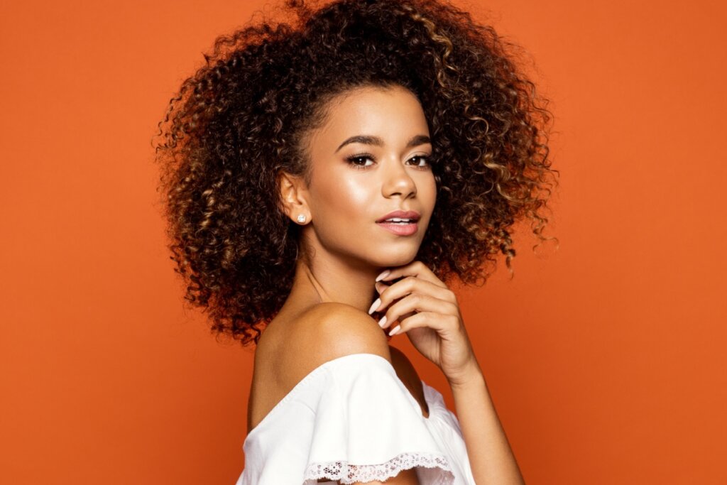 Mulher com cabelo cacheado curto com volume e usando blusa branca em fundo laranja
