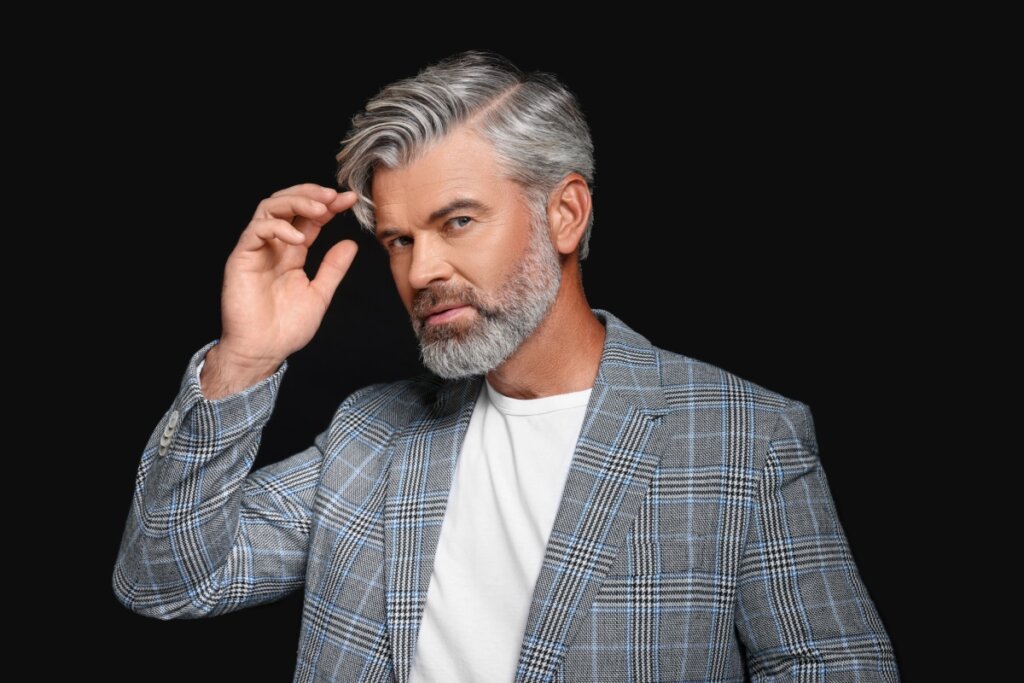 homem de cabelo e barba grisalho vestindo camiseta branca e blazer xadrez em fundo preto