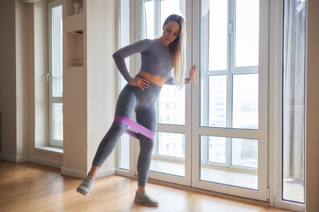 Mulher usando roupa cinza, com uma das mãos apoiadas em uma janela de vidro, fazendo abdução em pé com elástico roxo na perna 