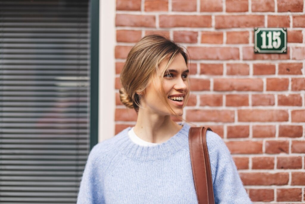 Mulher loira, com coque desconstruído, usando blusa azul, olhando para o lado e sorrindo