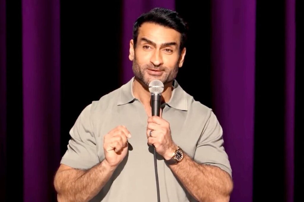 Ator e comediante Kumail Nanjiani em frente a um cortina roxa e segurando um microfone