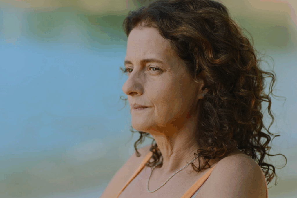 Cena mostrando uma mulher de perfil com expressão neutra diante de um fundo desfocado com água e luz natural