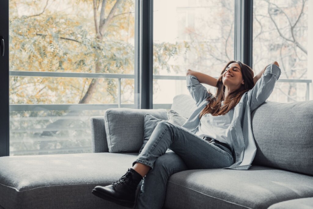 Mulher com o cabelo solto, usando camiseta branca e camisa listrada e calça jeans e bota preta sentada no sofá com as mãos atrás da cabeça descansando