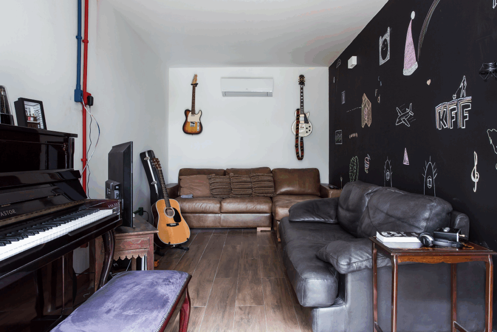 Sala de estar com sofás em marrom e preto, parede de lousa e com desenhos que remetem a música com guitarras penduradas na parede e violão em pedestal e piano ao lado de móvel com TV