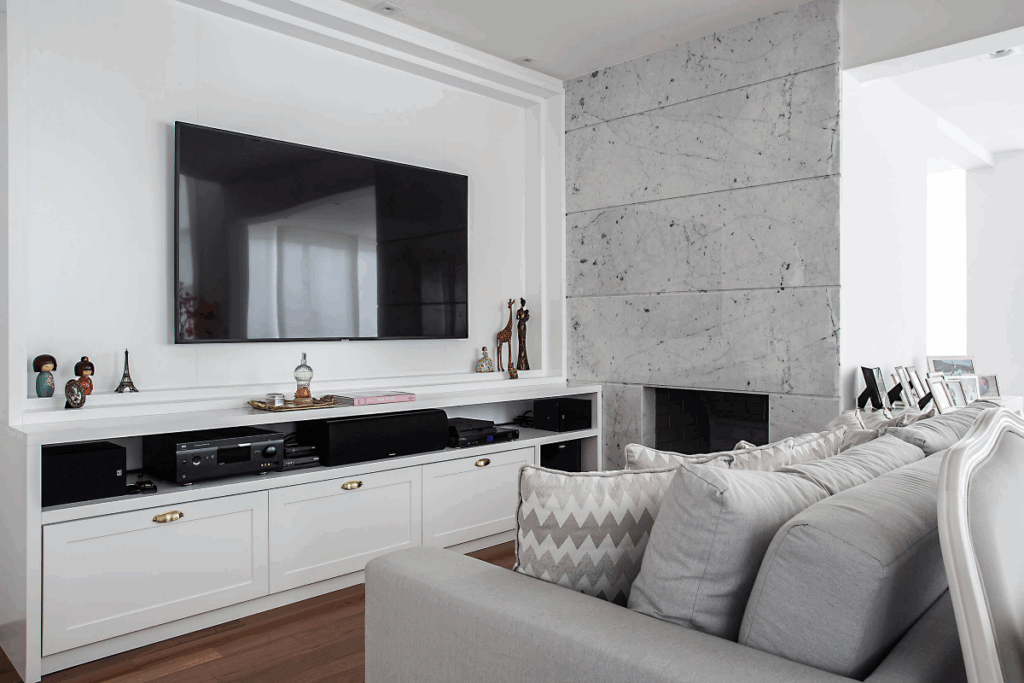 Sala de estar com TV na parede, as paredes em tons de branco e cinza-claro com móveis em branco com detalhe preto e sofá cinza-claro com almofadas estampadas em tons claros