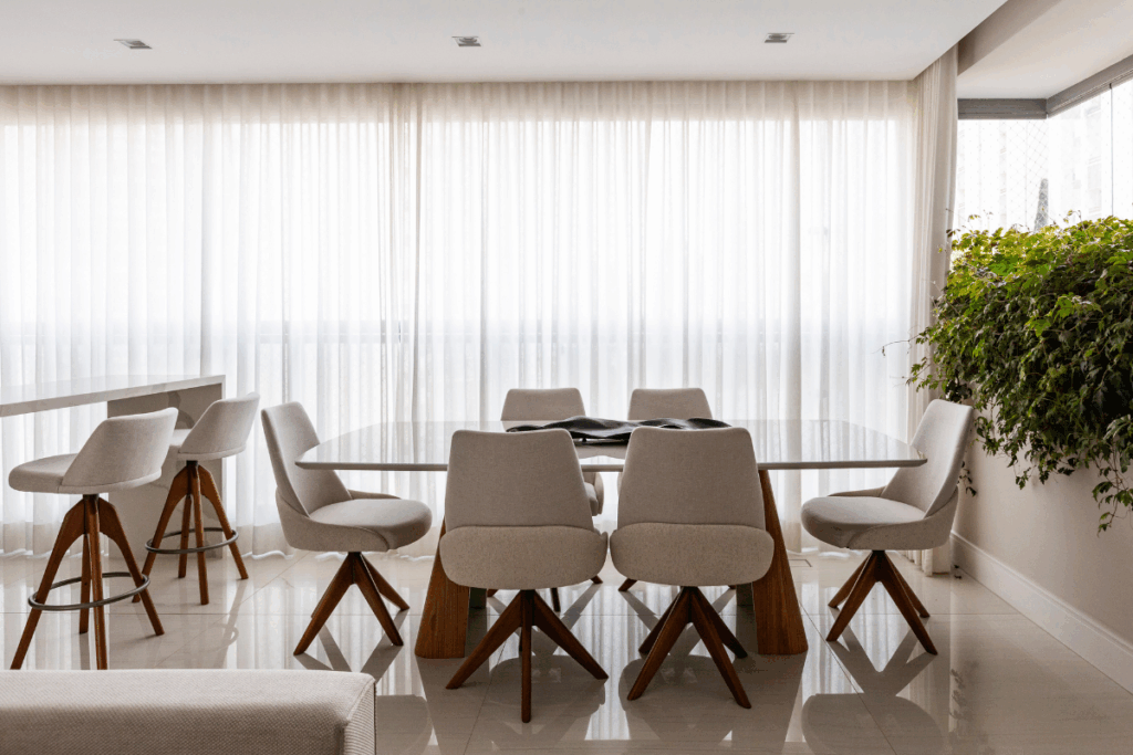 Sala de jantar moderna e iluminada com uma grande mesa de tampo claro e seis cadeiras estofadas em tom bege com pés de madeira. Ao fundo, uma ampla janela coberta por cortinas brancas translúcidas permite a entrada de luz suave. À direita, um maciço de folhagens verdes pendentes brota de um canteiro lateral, trazendo um elemento natural ao ambiente de tons neutros e piso brilhante.