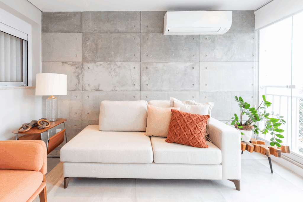 Sala de estar com parede de destaque em concreto aparente. Um sofá de dois lugares em tecido off-white é decorado com almofadas neutras e uma almofada terracota em tricô. À direita, sobre um banco de madeira rústica de formato irregular, há um vaso branco com uma planta Zamioculca. O ambiente possui iluminação natural vinda de uma varanda envidraçada lateral.