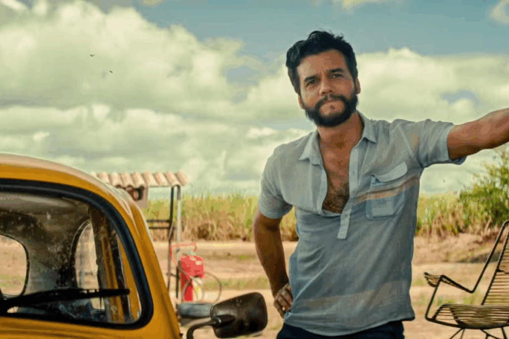 Personagem de Wagner Moura parado ao lado de um fusca amarelo em uma estrada árida, clima de interior