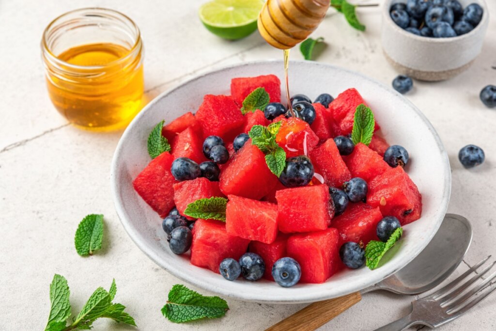 Salada de frutas com cubos de melancia, mirtilos, folhas de hortelã e mel sendo despejado em um recipiente branco