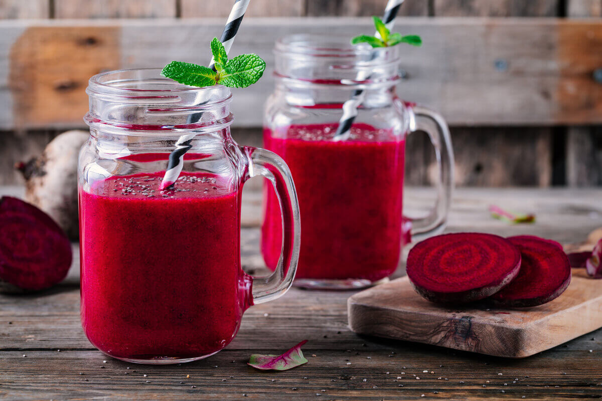 Dois copos de vidro com suco de beterraba em cima de uma mesa de madeira com uma tábua com rodelas de beterraba