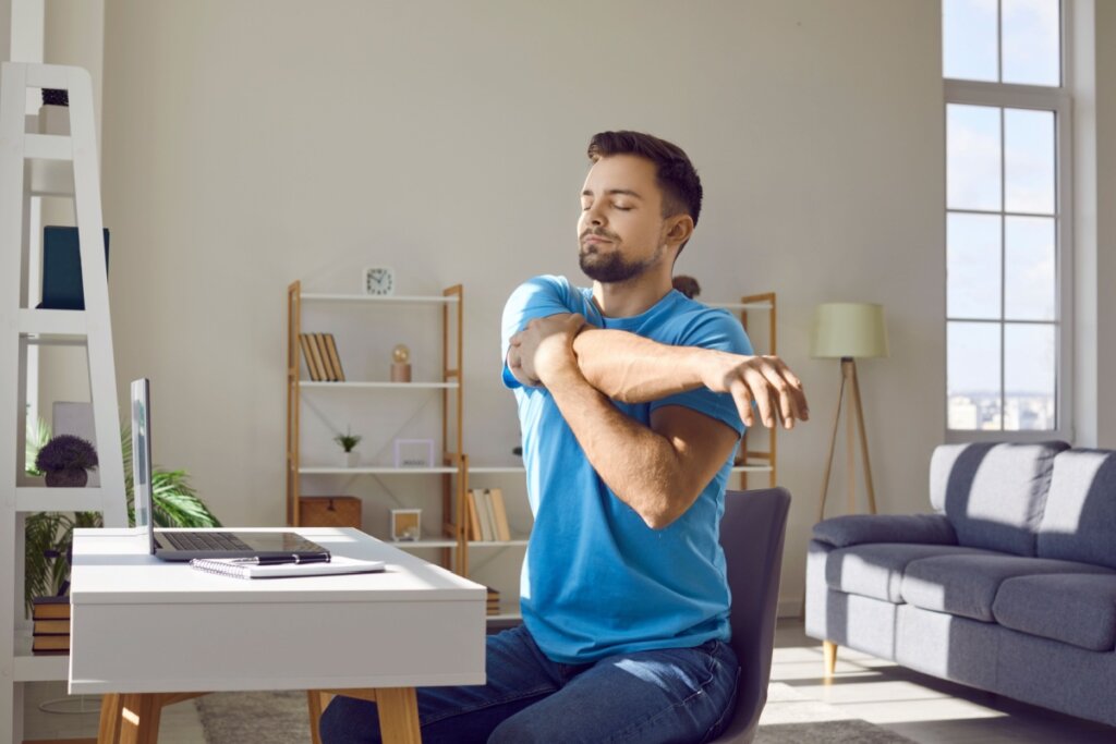 Homem com cabelo curto e barba usando camiseta azul, sentado em escritório em casa segurando um braço com o outro