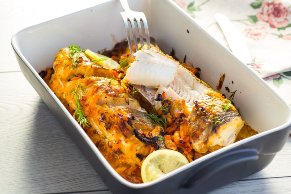 Peixe assado com cenoura, ervas e limão servido em refratário de cerâmica branca e azul em cima de mesa de madeira