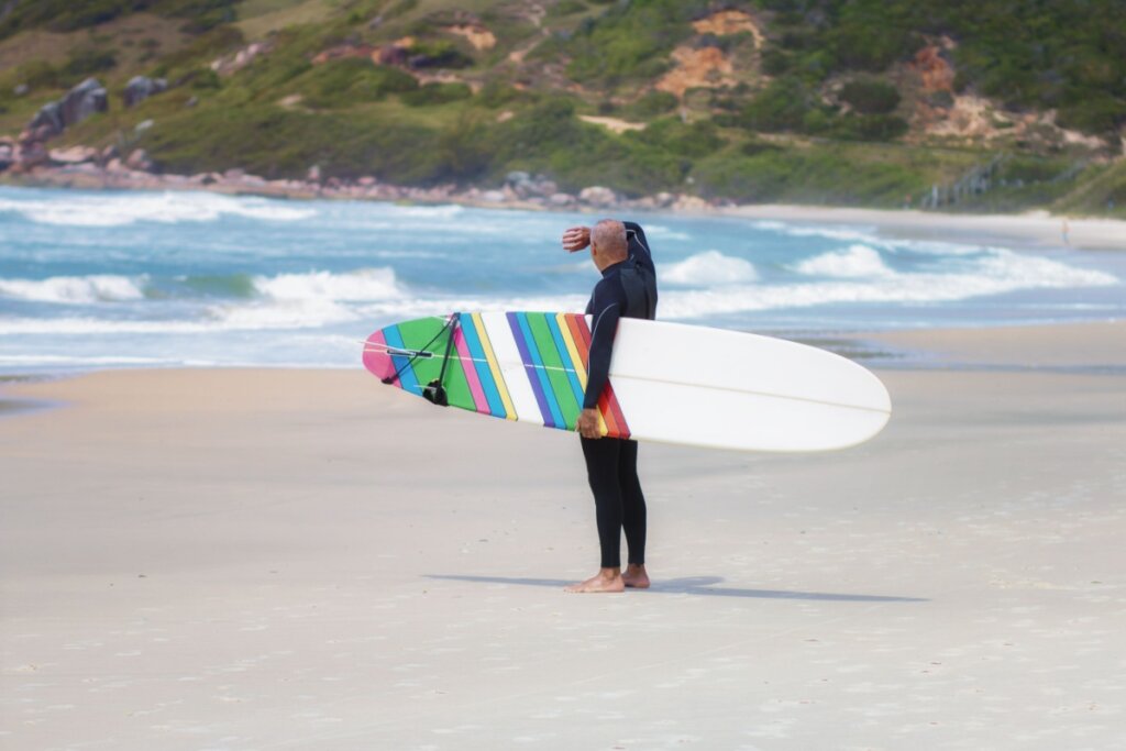 Um surfista de costas, usando roupa de neoprene preta, observa o mar na Praia do Rosa. Ele segura sob o braço uma prancha de surfe longa (longboard) branca com uma pintura colorida em listras verticais (arco-íris) na parte traseira. A areia é clara e o mar ao fundo apresenta ondas com espumas brancas. Ao longe, vê-se a encosta de um morro com vegetação verde e tons de terra.