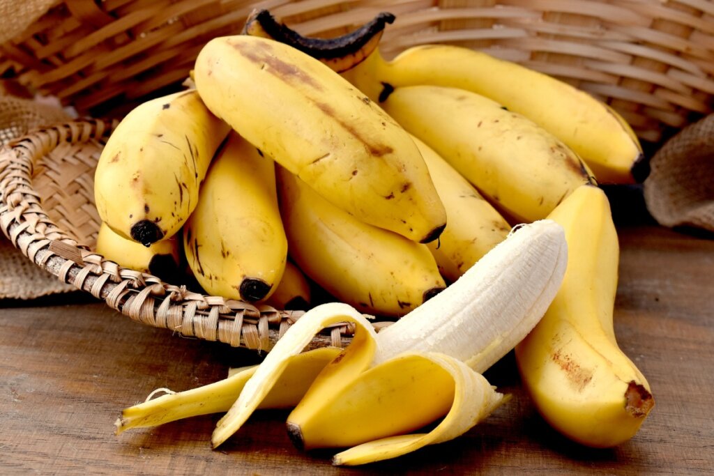 Cesto com várias bananas maduras e uma delas parcialmente descascada sobre mesa de madeira