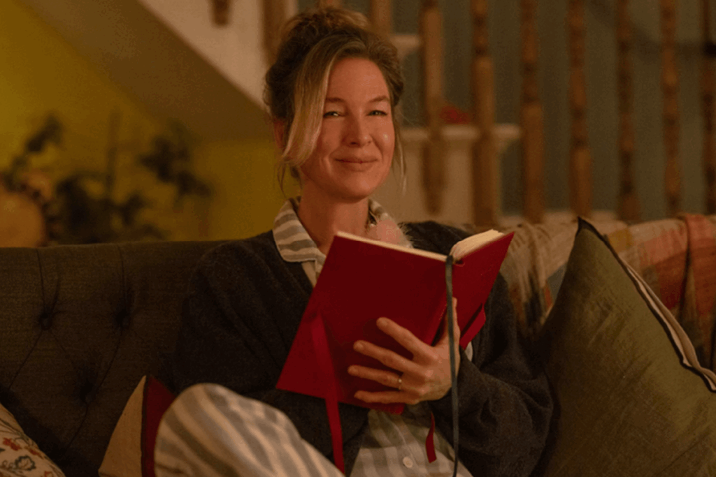 Mulher segurando um caderno vermelho e sorrindo enquanto está sentada no sofá em um ambiente aconchegant