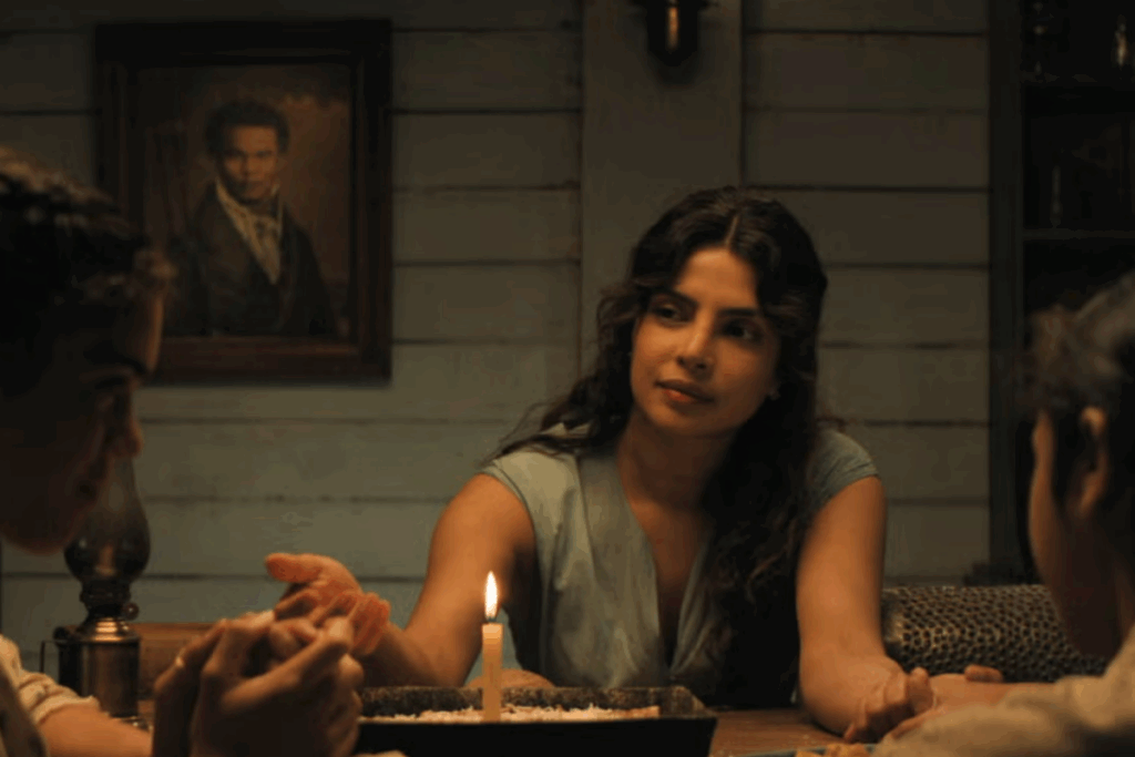 Imagem de mulher com o cabelo semi preso, usando regata cinza, de mãos dadas com duas crianças, todas sentadas à mesa