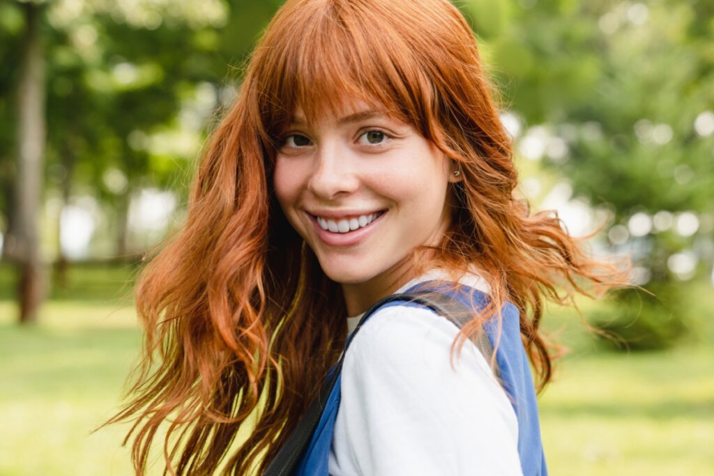 mulhjer ruiva de cabelo cacheado e franja sorrindo e de lado. árvores verdes desfocadas ao fundo