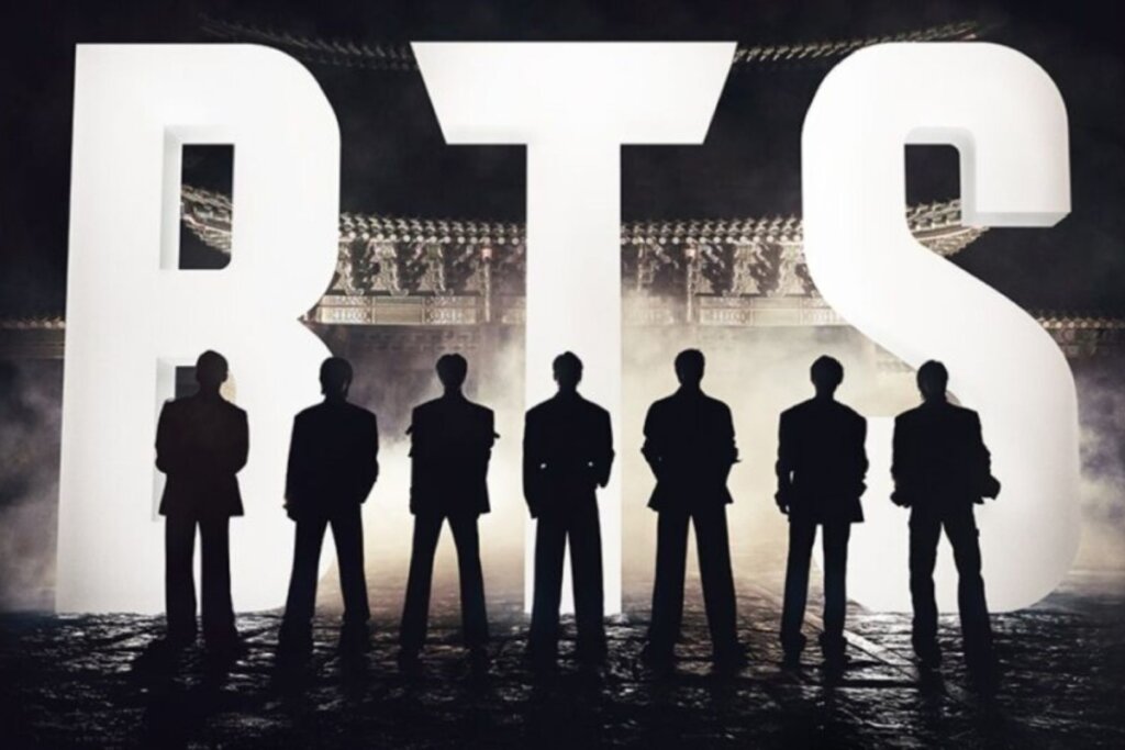 Silhueta de sete homens, os integrantes do grupo de K-pop BTS, vistos de costas e alinhados horizontalmente. Eles estão posicionados em frente a grandes letras brancas iluminadas que formam a sigla "BTS".